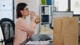 Businesswoman eating bite of tasty sandwich drinking coffee in front of monitor during takeout lunchtime. Fast food order paper bag delivered. Woman analyzing management strategy
