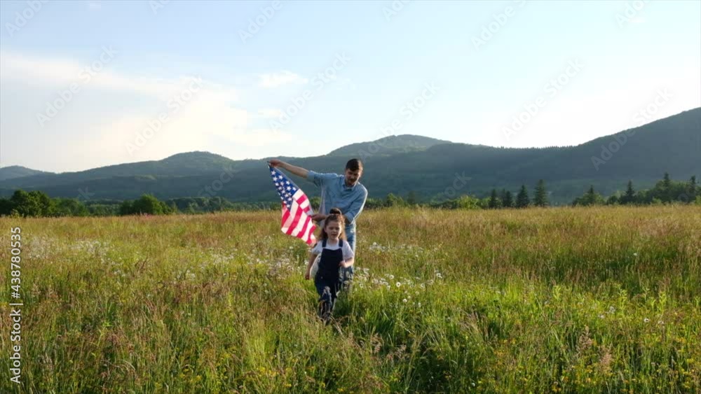 Caucasian pretty joyful little daughter and young bearded father runs with USA flag on background summer mountains. Positive family with American flag as symbol of freedom, youth and independence Caucasian pretty joyful little daughter and young bearded father runs with USA flag on background summer mountains. Positive family with American flag as symbol of freedom, youth and independence