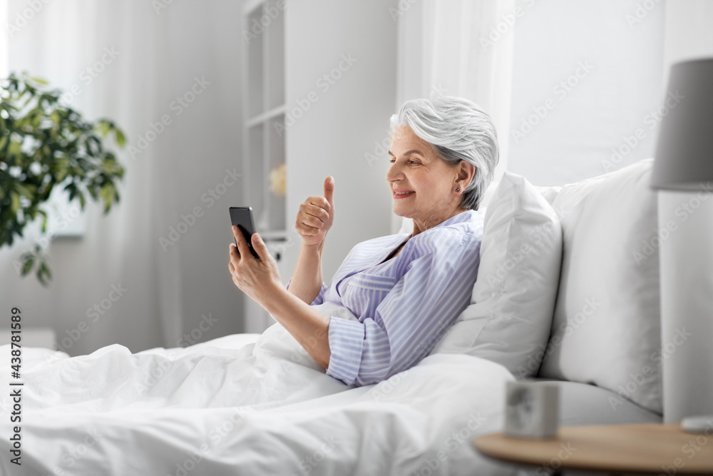 old age, technology and people concept - happy smiling senior woman in pajamas using smartphone sitting in bed and having video call and showing thumbs up at home bedroom old age, technology and people concept - happy smiling senior woman in pajamas using smartphone sitting in bed and having video call and showing thumbs up at home bedroom