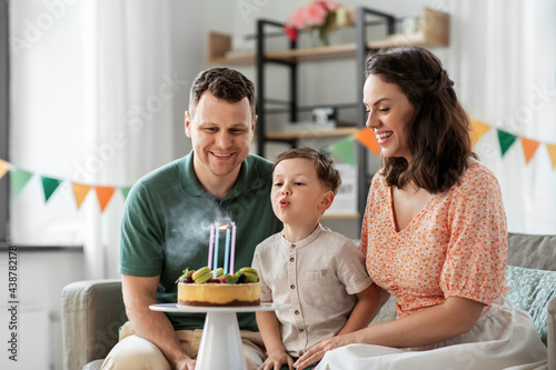 family, holidays and people concept - portrait of happy mother, father and little son blowing to four candles burning on birthday cake sitting on sofa at home party