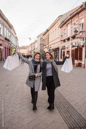Two beautiful fashion Caucasian women walk down the street smiling and carrying bags in their hands. Autumn shopping in the city