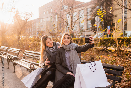 Summer sunny day, two young laughing women sitting on a park bench, rest after shopping and using their smartphones. Nearby are shopping bags. Two friends sitting in park and taking selfie.