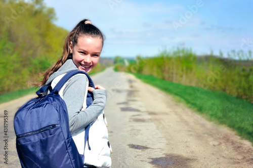 A young girl with a blue backpack turned around and smiling looks at the camera squinting at the sun against the background of a blue sky, stretching into the distance of the road plants and trees.