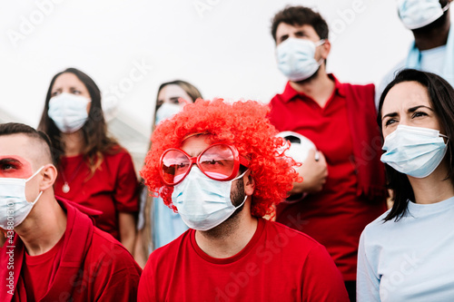 Group of football team supporters in red and blue t-shirt and protective face mask watching concentrated the match from the fan zone at stadium - Live sports new normality concept photo