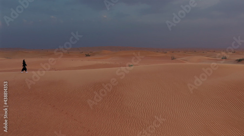 AERIAL. Camera following woman in traditional Emirati dress walking in a desert in strog wind and sunset.