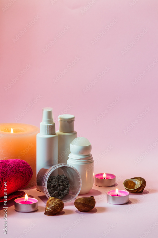 Hygiene products in white bottles, an alginate mask and a pink towel stand against a pink background next to burning candles. Hygiene products in white bottles, an alginate mask and a pink towel stand against a pink background next to burning candles.