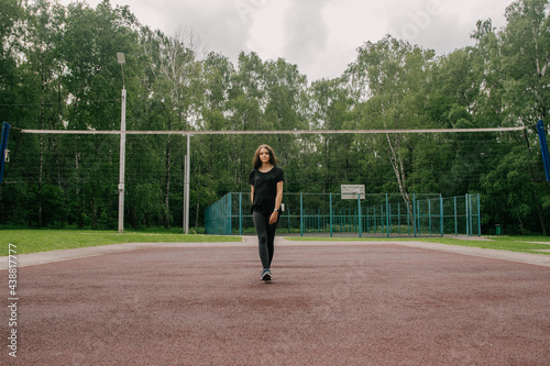 a girl in a black T-shirt and leggings stands on the sports ground near the net for playing volleyball © Наталья Анюхина