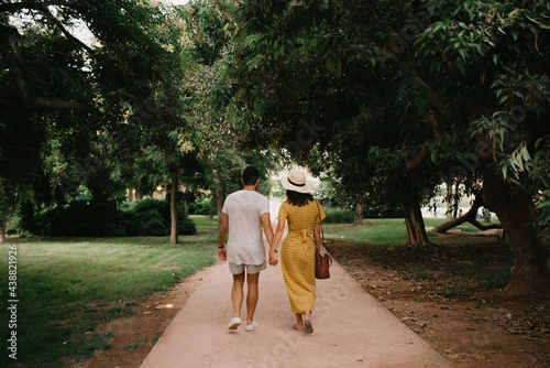 A photo from behind of a brunette girl in a yellow dress and her boyfriend who are walking on a sand path between trees in the Valencian park. A couple of tourists on a date in the evening.