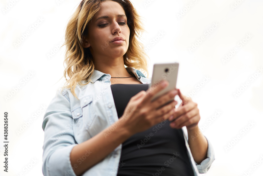 Below view of millennial blogger 20s using modern cellular device for online messaging, beautiful hipster girl reading text publication while browsing media content in mobile social networks Below view of millennial blogger 20s using modern cellular device for online messaging, beautiful hipster girl reading text publication while browsing media content in mobile social networks