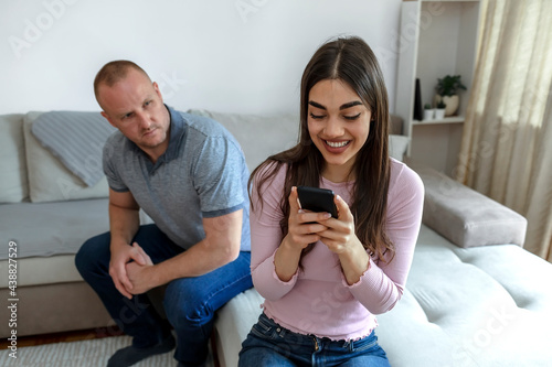Jealous curious man spying his girlfriend's smartphone while she is typing message to her lover and smiling happily. Jealous husband spying the phone of his partner. Man spying his girlfriend mobile. photo