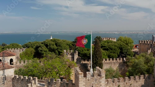 Close-up of national and municipal flags on poles over medieval stone Saint George Castle. Drone camera flying backward away from landmark. Lisbon, capital of Portugal.