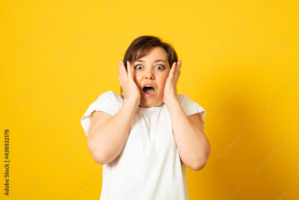 Photo of scared pretty woman keeps hands on cheeks, looks with surprisement and fear at camera, wears casual white t-shirt, isolated over yellow background. Omg concept.