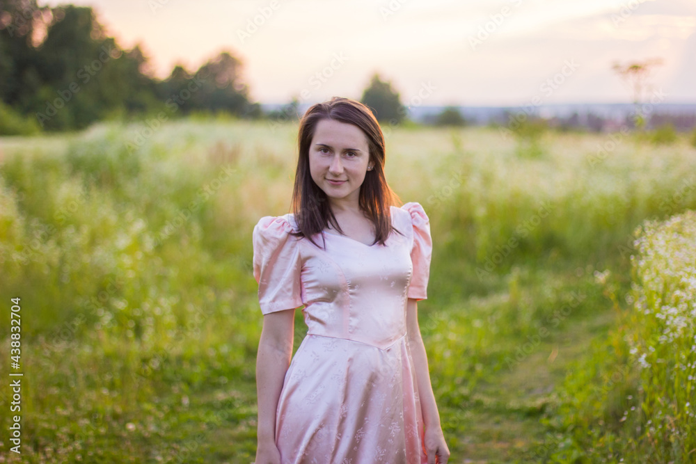 Beautiful portret of happy woman with dark hair standing in the wildflowers field holding the bottom of the long pink dress on a warm summer evening. Joyfull summer walking. Beautiful portret of happy woman with dark hair standing in the wildflowers field holding the bottom of the long pink dress on a warm summer evening. Joyfull summer walking.