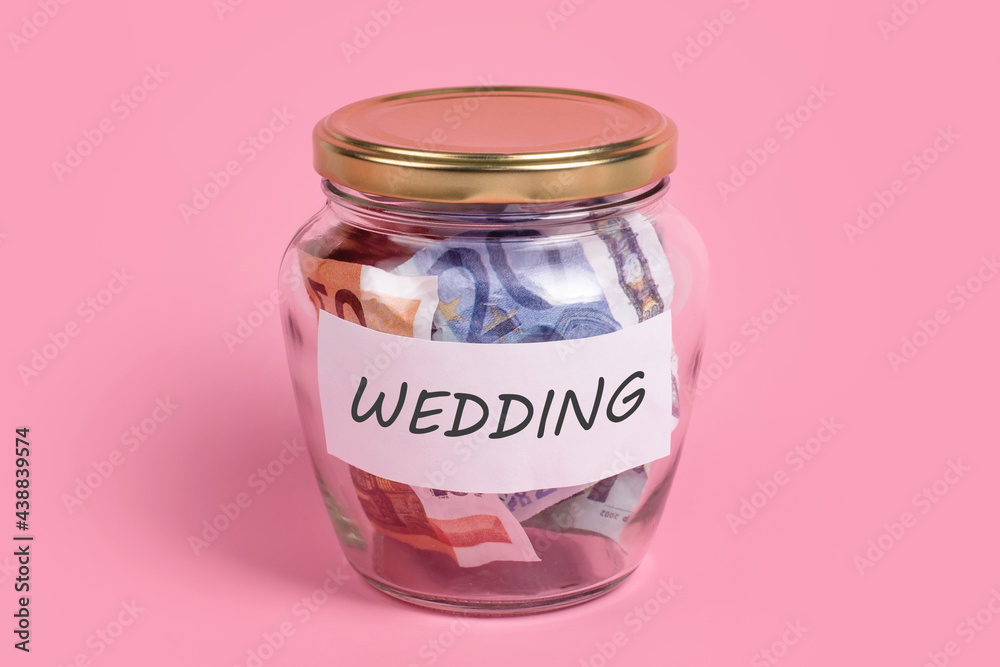 Money box with word WEDDING on sticky note paper. Euro banknotes in glass money jar with savings label, financial, saving. Jar full of cash, save money concept, expense control, future planning.