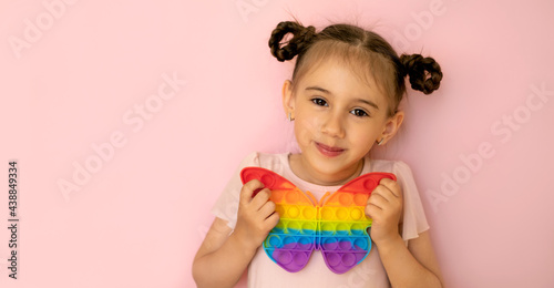 A beautiful cute little girl on a pink background holds a popit toy in the shape of a butterfly in her hand. Funny pigtails for a girl . Children s entertainment and educational toys pop it. 