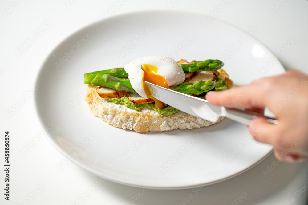 Hand holding knife and cutting poached egg on delicious morning toast with cream cheese, mashed avocado, roasted chicken slices, asparagus on white plate. Healthy nutritious breakfast recipe. 