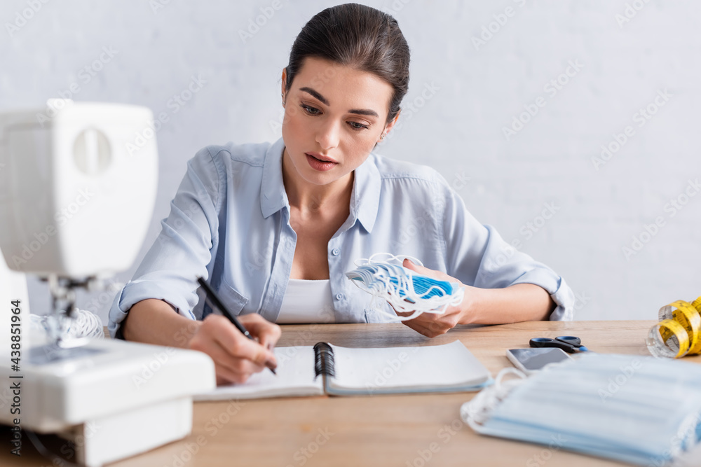 Seamstress writing on notebook near medical masks and blurred sewing machine