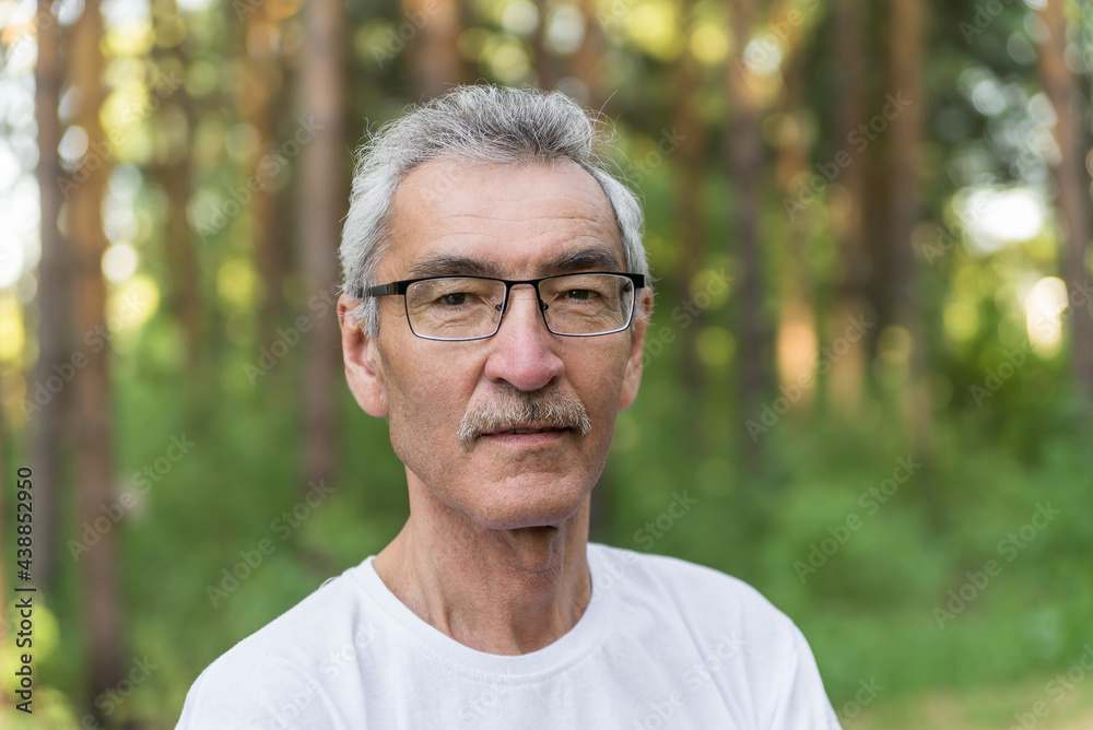 portrait of an elderly man on the background of the forest park. an old man of 60s against the background of trees wearing glasses and a white T-shirt portrait of an elderly man on the background of the forest park. an old man of 60s against the background of trees wearing glasses and a white T-shirt