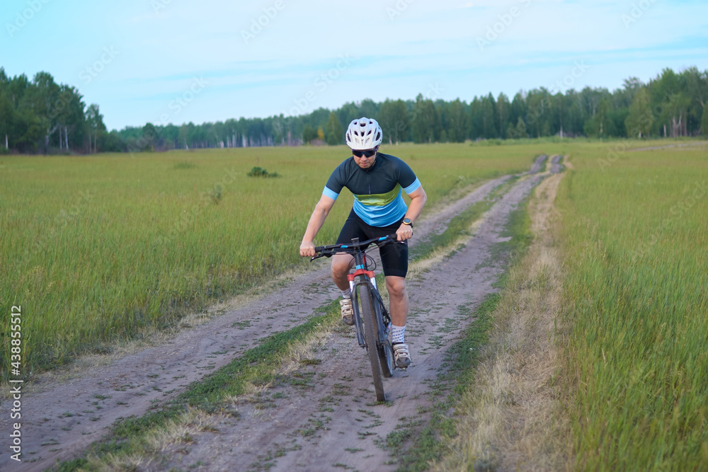 A male cyclist quickly rides a mountain bike on a dirt road in a field. Dressed in a bicycle uniform, helmet and sunglasses. HIIT workout. Outdoor sports and recreation. Healthy lifestyle.