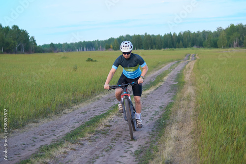 A male cyclist quickly rides a mountain bike on a dirt road in a field. Dressed in a bicycle uniform, helmet and sunglasses. HIIT workout. Outdoor sports and recreation. Healthy lifestyle.