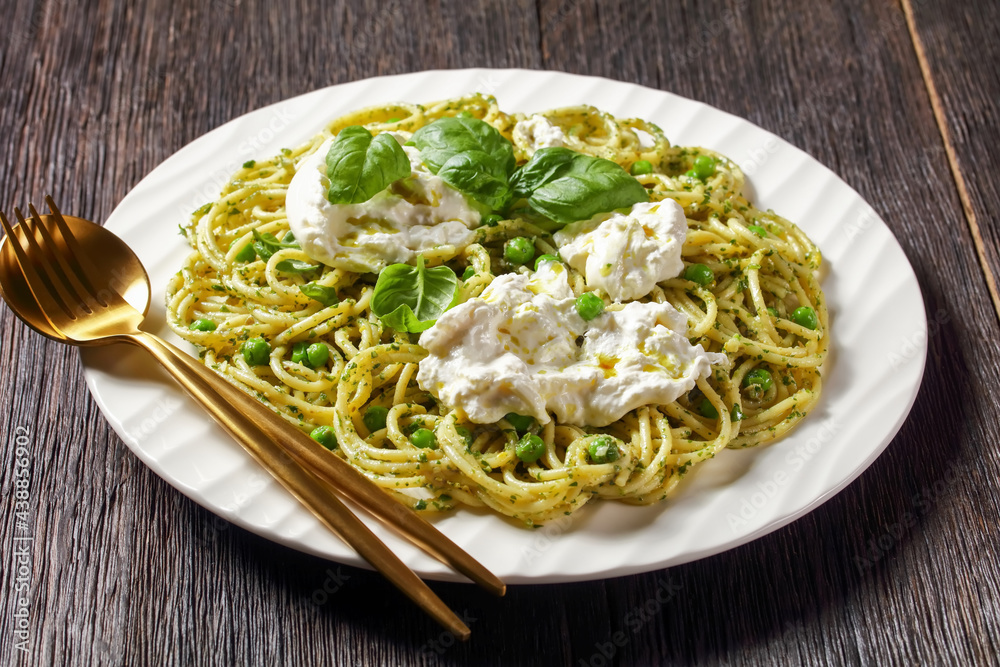 pesto spaghetti with green peas topped with torn mozzarella ball, fresh basil leaves on a plate, italian cuisine