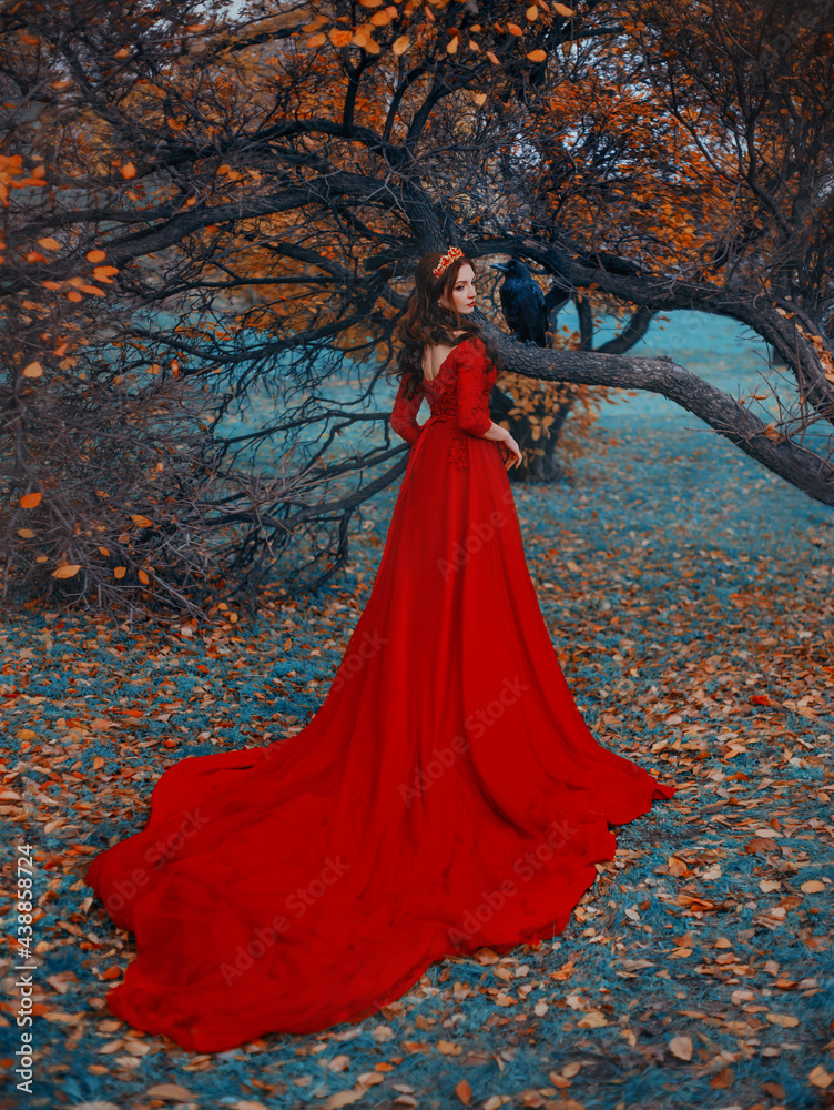 Fantasy woman queen in red dress with long train. Golden crown on head. Autumn nature tree black raven sits on branch, fallen orange leaves on green grass blue fog. rear view back. Girl gothic queen.