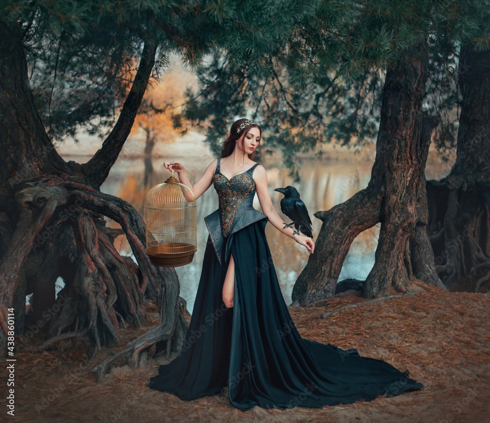 Fantasy woman witch in creative black gothic dress. Raven sitting on hand.  queen holds golden cage. Nature forest river. fashion model fantasy outfit.  Dark woods trees mystical nature. Girl princess Stock Photo
