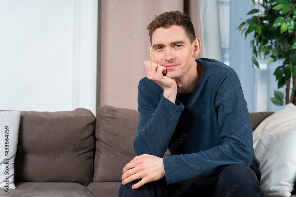 Portrait of handsome happy positive guy, young bored happy man sitting on cozy couch or sofa at home props his chin with hand in pyjamas, looking at camera and smiling. Portrait of handsome happy positive guy, young bored happy man sitting on cozy couch or sofa at home props his chin with hand in pyjamas, looking at camera and smiling.