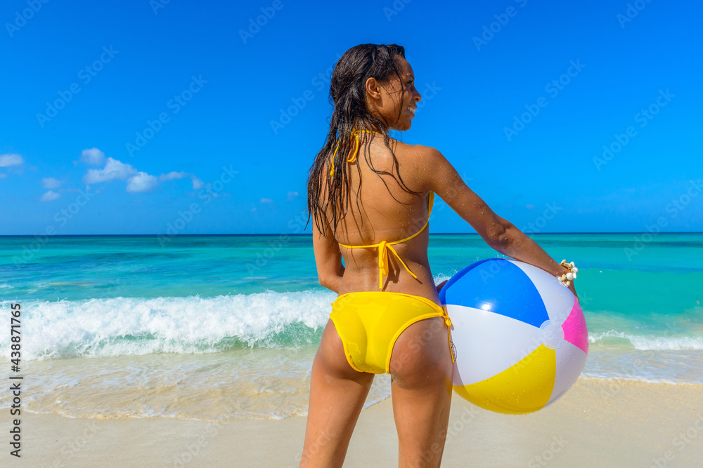 Young beautiful slim, interracial, black woman standing on the beach with a yellow bikini and beach ball