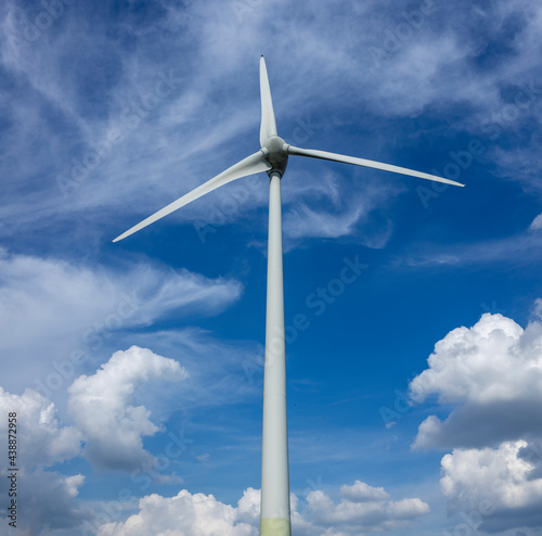 Square high resolution frame of tall wind electricity turbine mill with blades rising up among fluffy and whispery clouds against a vibrant blue sky. Sustainable energy concept. 