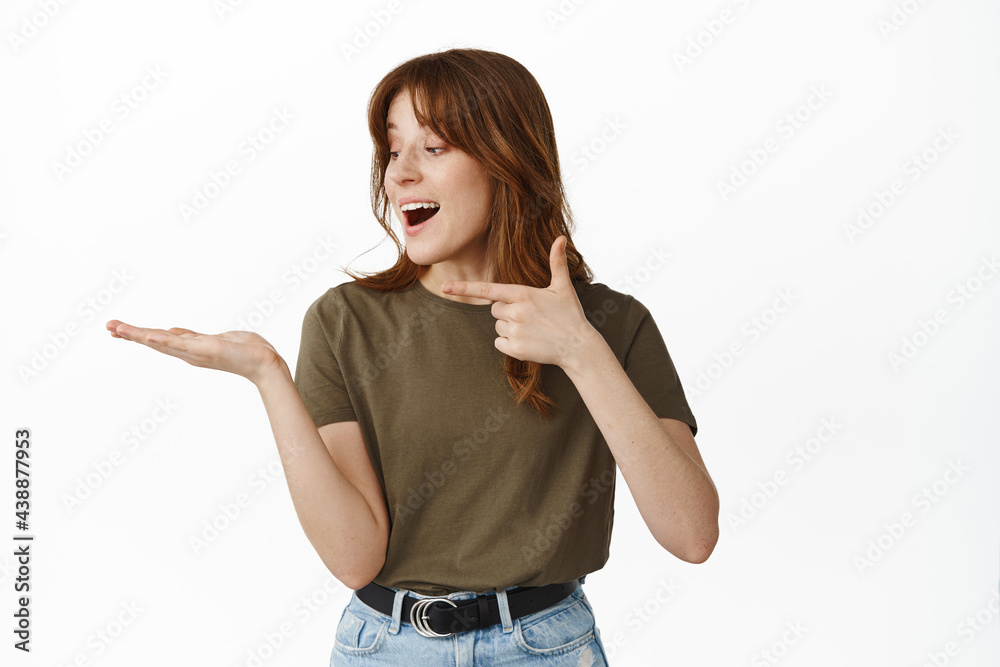 Amazed and happy young woman, pointing and looking at her open hand holding object, showing item on palm against empty copy space, standing over studio background Amazed and happy young woman, pointing and looking at her open hand holding object, showing item on palm against empty copy space, standing over studio background