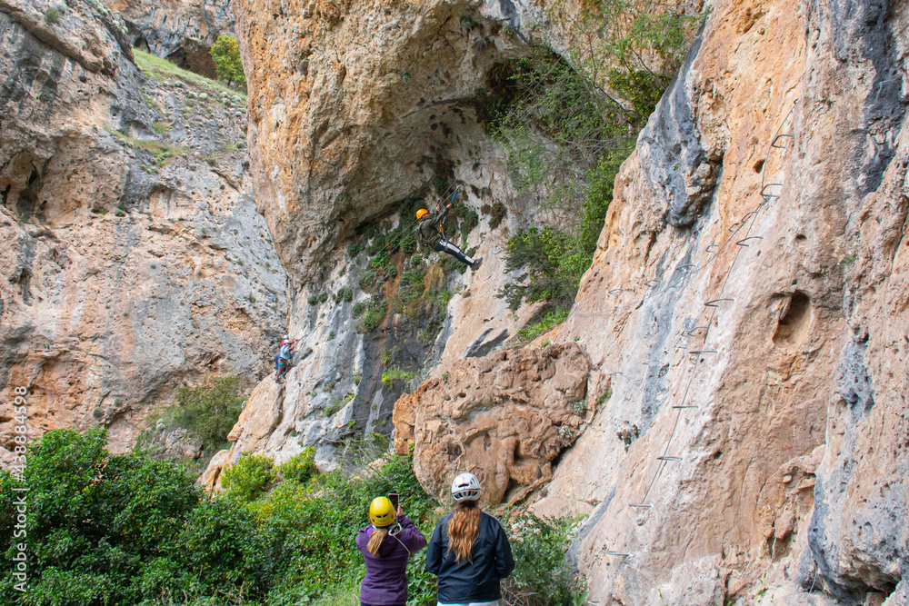 A woman watching and another taking a photo of a friend while riding a zip line on a via ferrata