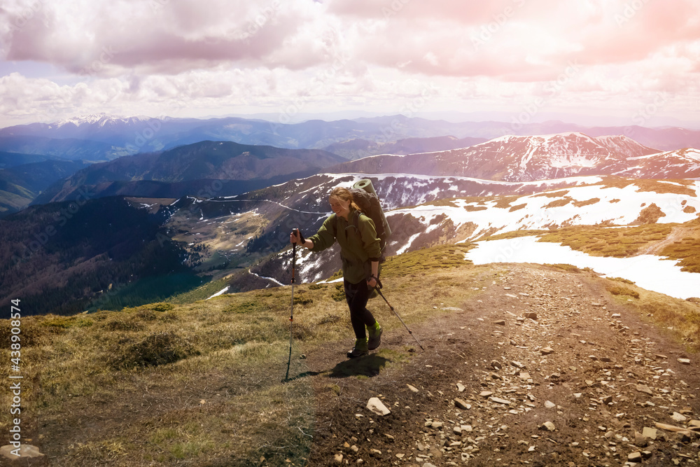 Obraz premium Young smiling girl climbs the mountains with trekking poles and a backpack in early spring, on the background there is a beautiful view with snowy mountains. Traveler goes hiking in the national park.