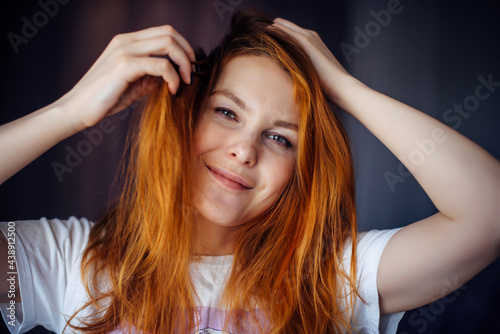 Portrait of beautiful young woman with thick, tousled red hair, close-up. Green-eyed beauty touches her hair while looking at camera and smiling. Advertising of care, hair coloring.