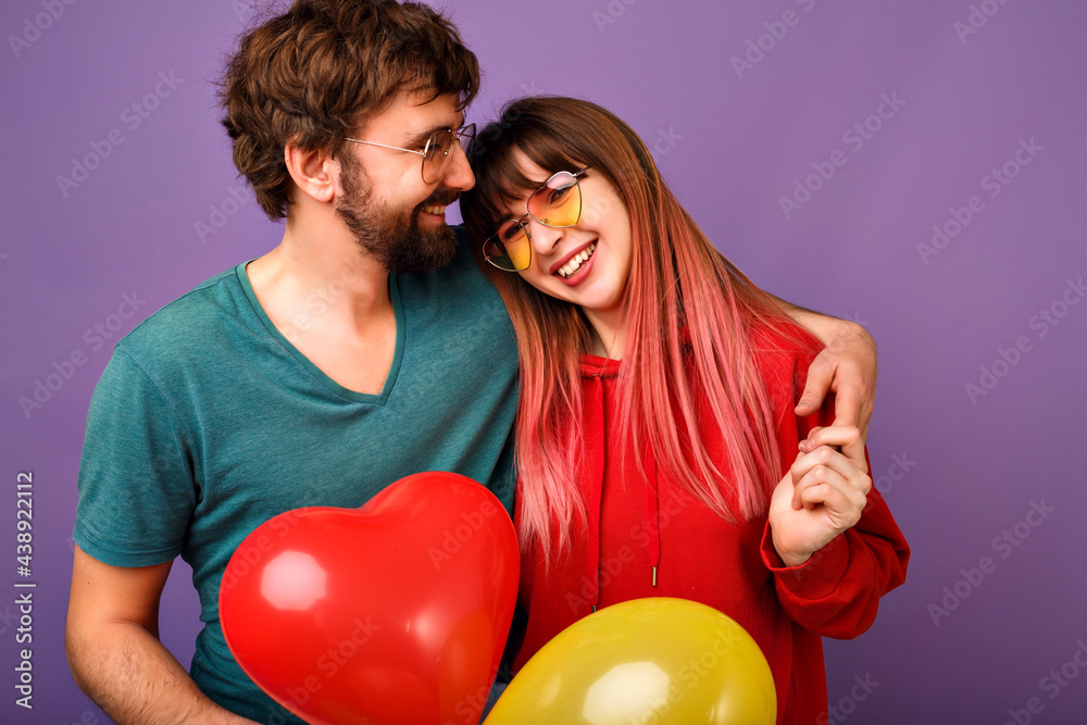 Young loveble hipster couple posing on violet background, bright trendy casual clothes and glasses, hugs and smiling, friendship and relation goals, holding air balloons, ready for celebration. Young loveble hipster couple posing on violet background, bright trendy casual clothes and glasses, hugs and smiling, friendship and relation goals, holding air balloons, ready for celebration.
