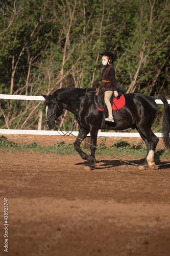 Young girl riding black horse wearing surgical mask 