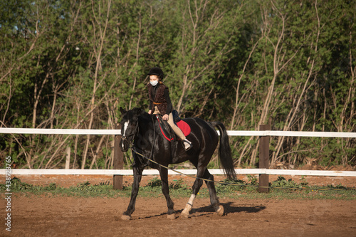 Young girl riding black horse wearing surgical mask 