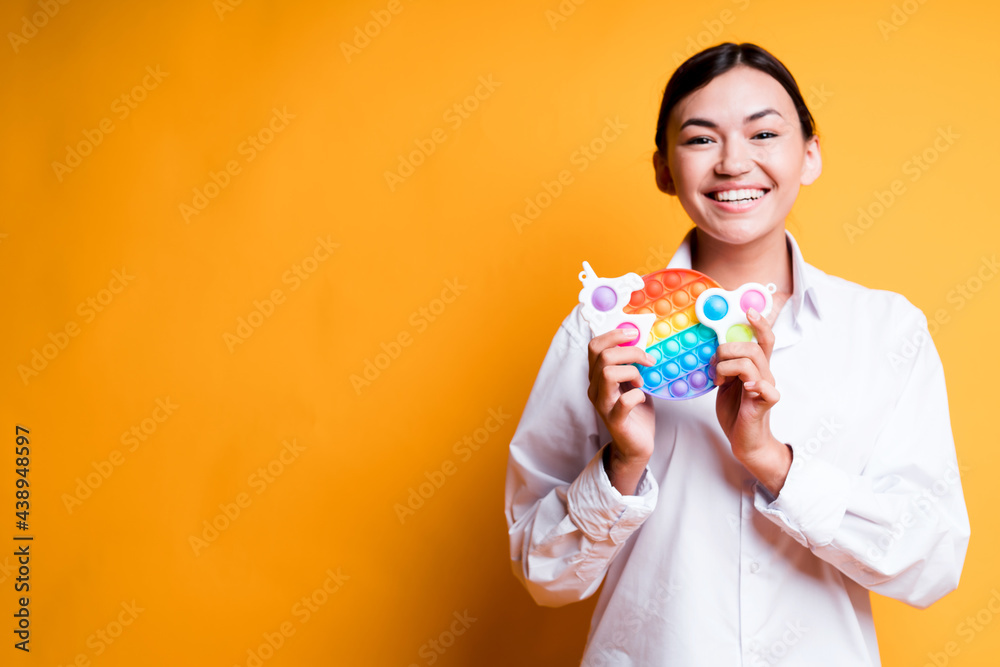 Happy asian girl in shirt holding childrens toys pop it and simple dimple smiling broadly while standing on yellow background Happy asian girl in shirt holding childrens toys pop it and simple dimple smiling broadly while standing on yellow background