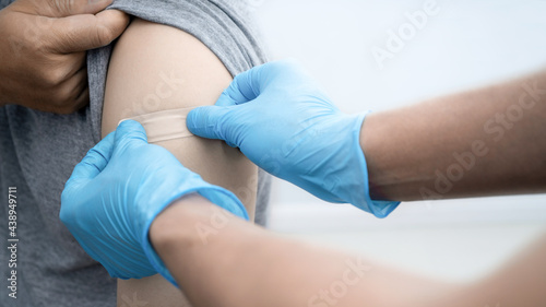 Doctor in blue rubble PPE protective gloves putting adhesive bandage plaster medical on man'arm after injection of COVID'19 CORONA Vaccine. © Yellow Boat