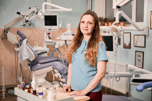 Beautiful young woman dentist looking at camera and smiling while standing by the table with instruments and tools in dental office. Concept of dentistry and dental staff.