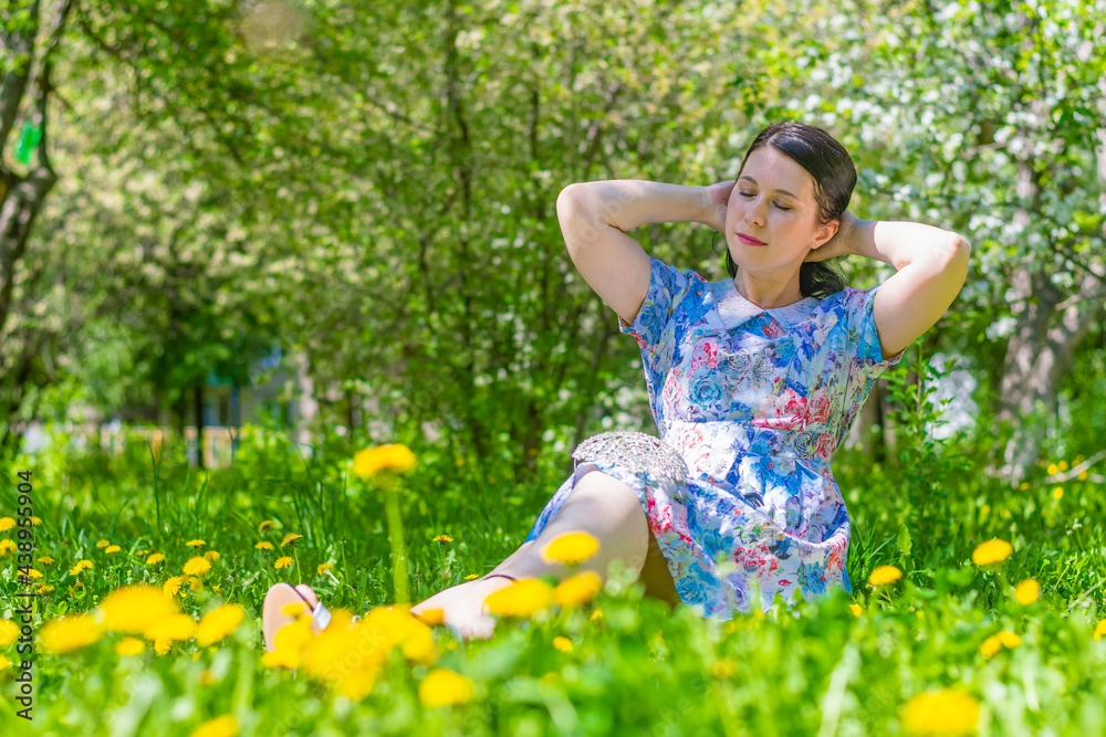 A young beautiful brunette girl in a summer blue dress sits on a lawn covered with yellow dandelions against a background of blooming apple trees. Straightens hair. A young beautiful brunette girl in a summer blue dress sits on a lawn covered with yellow dandelions against a background of blooming apple trees. Straightens hair.