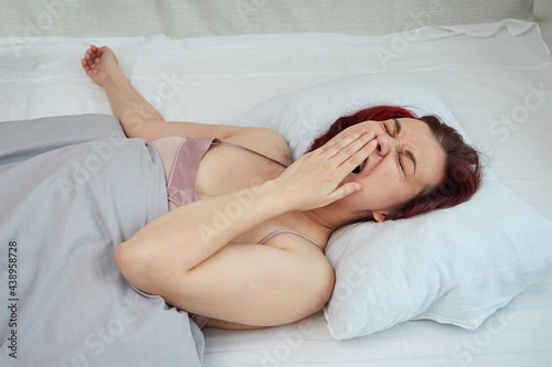 Young woman in pink top yawning covering her mouth with hand while lying in her bed