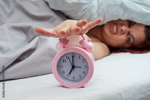 Displeased woman lying in bed stopping alarm clock in the early morning