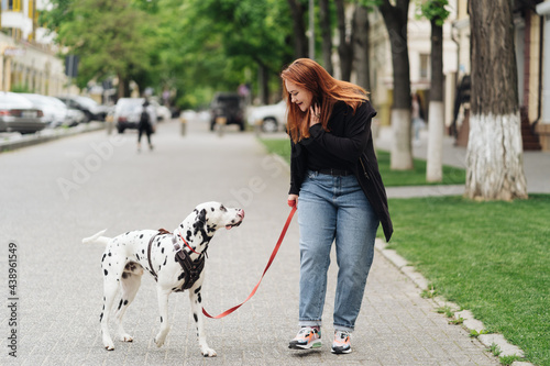 View of young Caucasian female cynologist walking at city urbanity during morning time for training dalmatian dog, plus size female spending leisure with pet best friend enjoying weekend at street