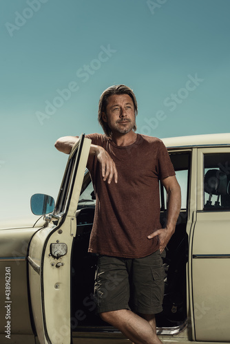 Blond man in brown t-shirt and shorts standing by open door of a classic car on a sunny day in the countryside.