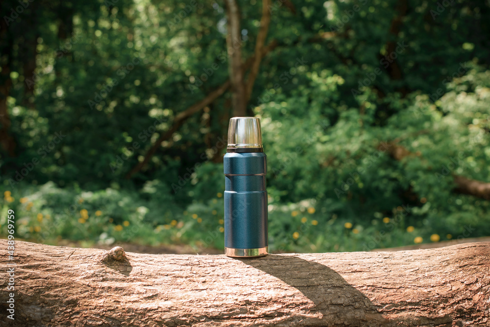 Traveling blue thermos on a log in the forest. Outdoor camping reusable flask. Concept of hiking, camping in nature Traveling blue thermos on a log in the forest. Outdoor camping reusable flask. Concept of hiking, camping in nature