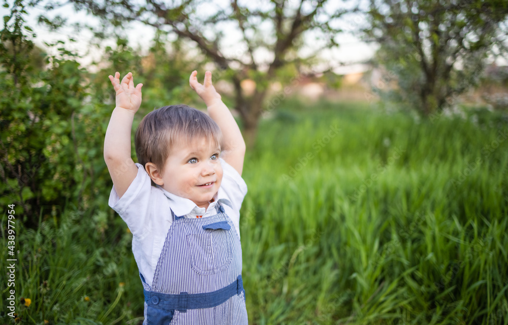 Cute boy in fashionable clothes with blue eyes plays and jumps in the tall grass in a large green blooming garden Cute boy in fashionable clothes with blue eyes plays and jumps in the tall grass in a large green blooming garden