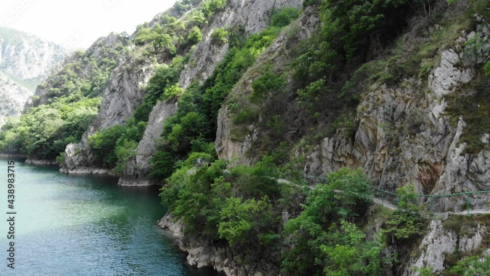 Drone view of Matka Canyon. Drone shot of a lake in a canyon in North Macedonia. Rocky green slopes. Transparent water surface of the lake. Mountain trail along the river. Lake in the mountains