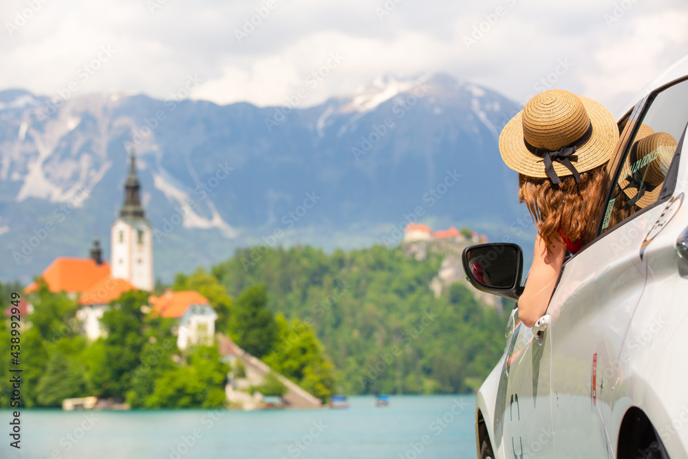 Happy young woman on vacation leaning out of car window on shore of Bled lake, country of Slovenia. Travel, holiday, tourism, explore, rent a car concept.