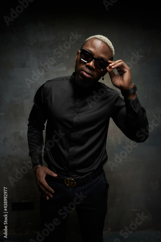 Elegant stylish handsome african man in black silk shirt, pants and sunglasses standing and posing against grey concrete wall